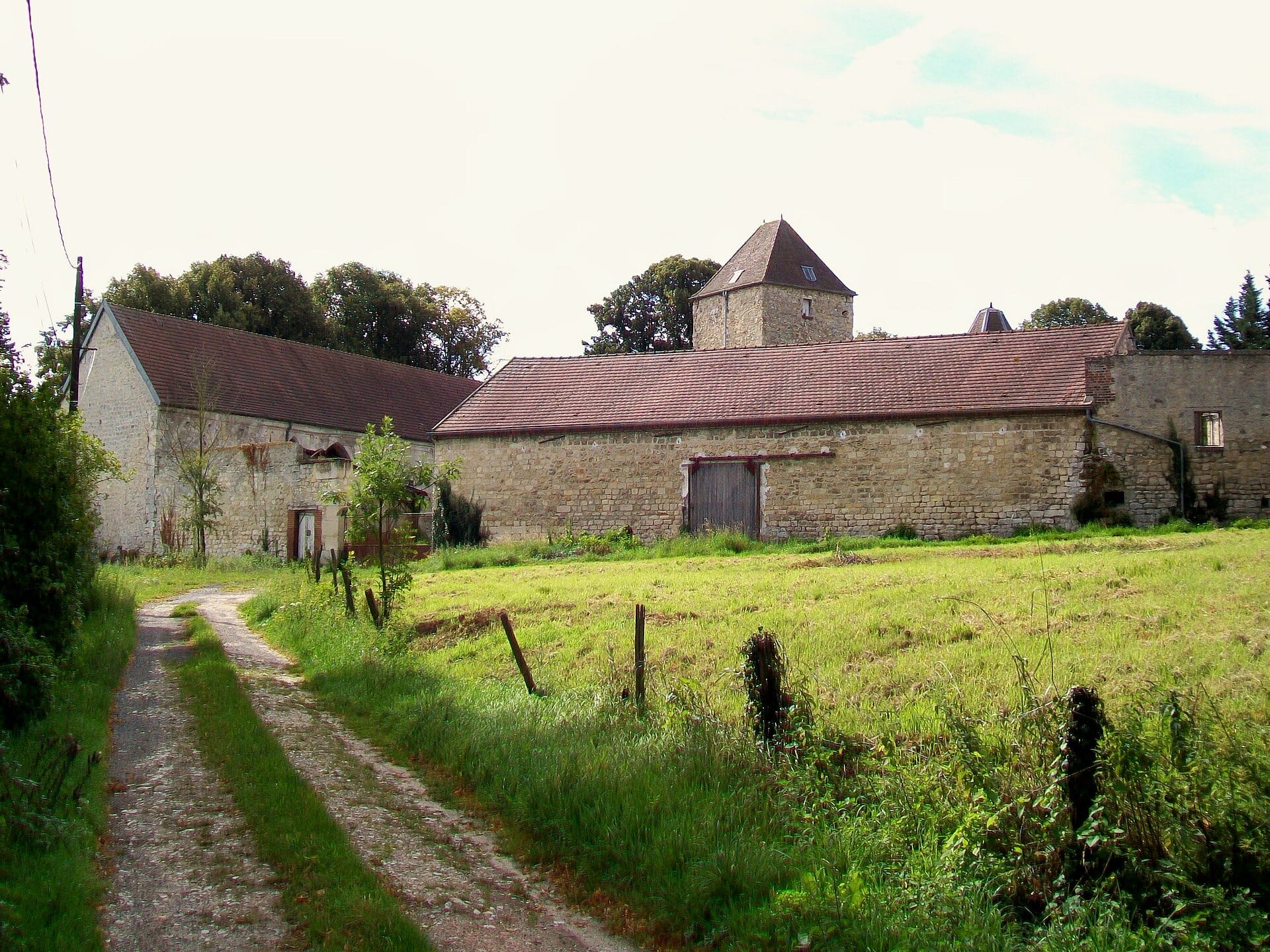 Longue ferme en pierre calcaire présentant vers l’extérieur de hauts murs presque aveugles, une large porte charretière en bois et une tour carrée coiffée d’un toit en pavillon dépassant les toitures de tuiles