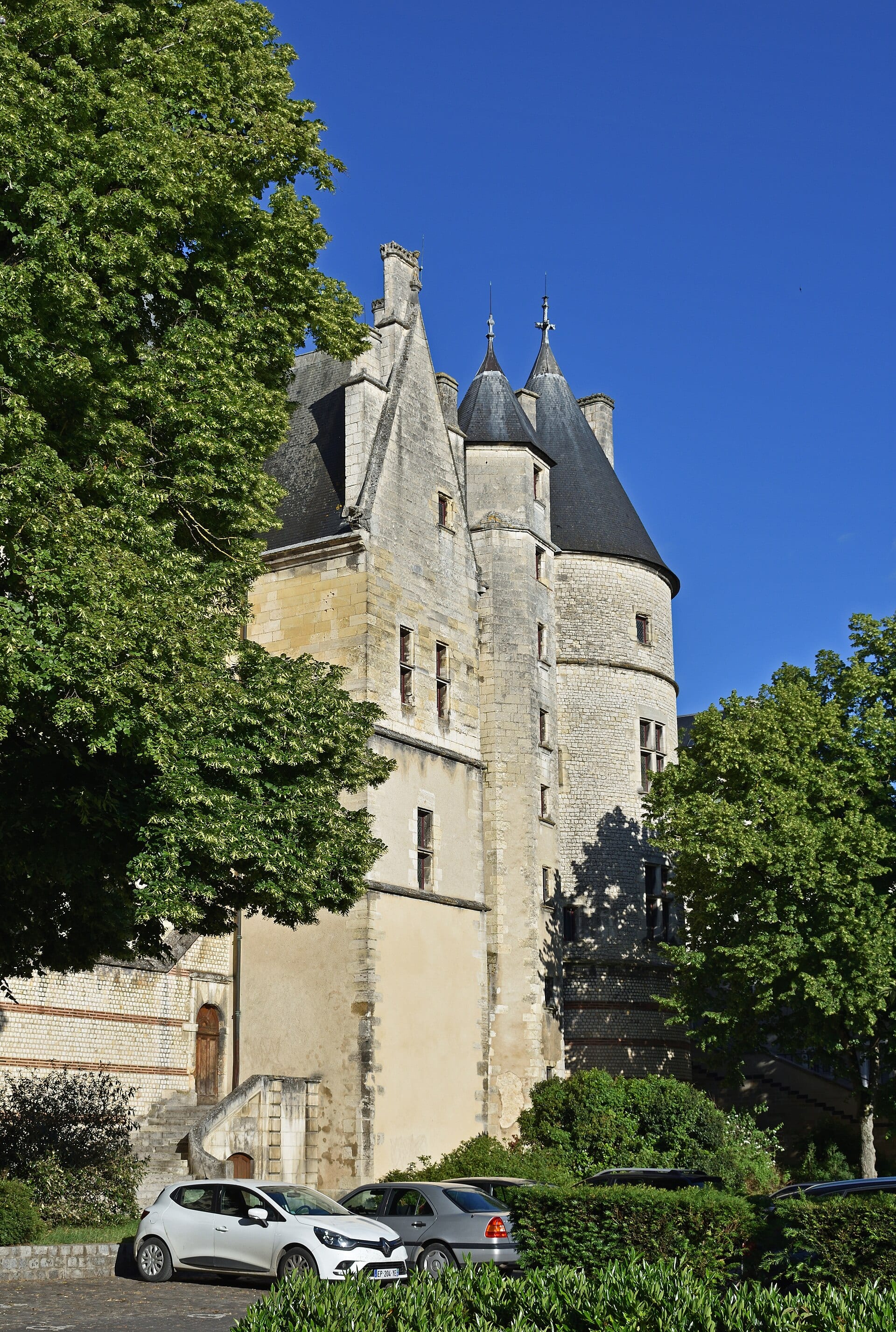 Le palais Jacques-Cœur de Bourges : haute tour d’escalier polygonale, toitures d’ardoise à lucarnes et tourelles en poivrière, murs de pierre claire, arbres et voitures au pied