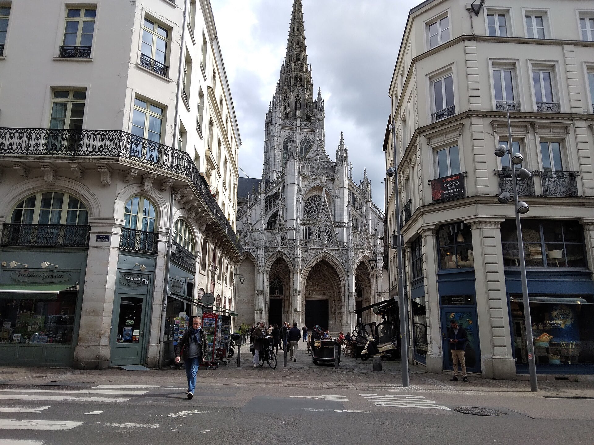 L’église Saint-Maclou de Rouen vue depuis une rue : façade à porches sous des gâbles ajourés, remplages, flèche de pierre ; immeubles et passants au premier plan