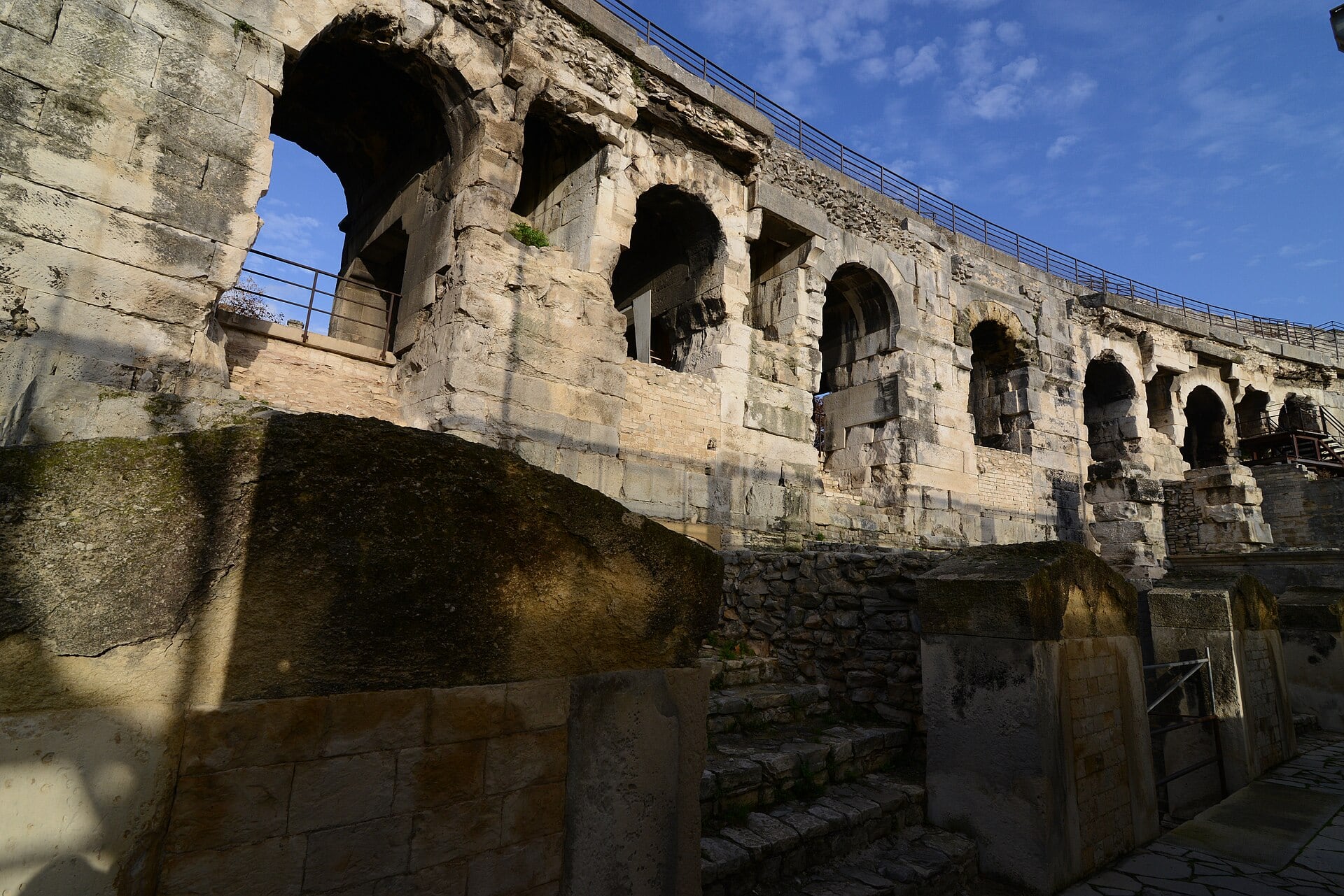 Portion du mur extérieur des arènes de Nîmes vue en contre-plongée : arcades superposées en plein cintre, appareil de pierre de taille, garde-corps au sommet
