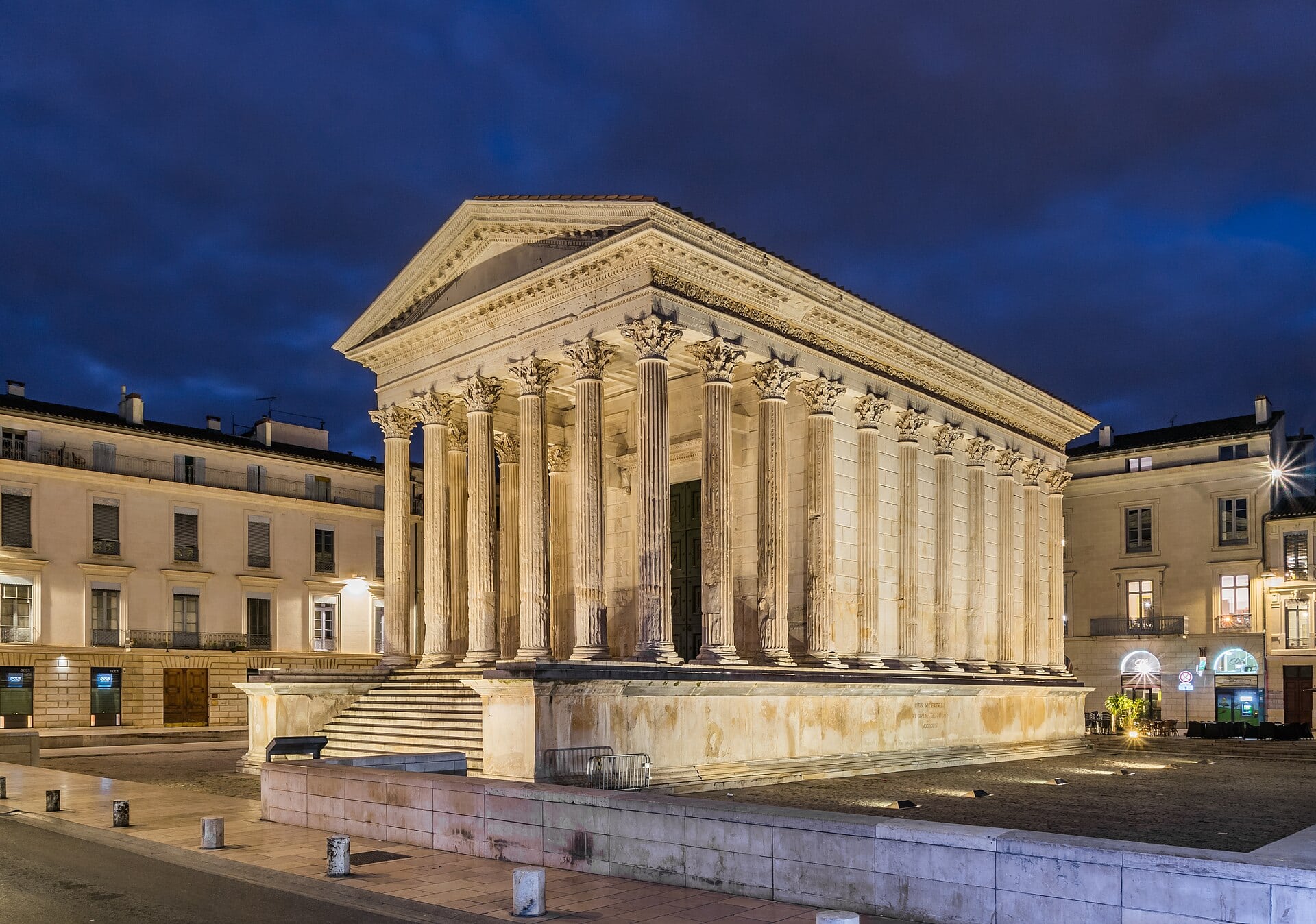 La Maison Carrée de Nîmes illuminée à la tombée du jour : temple romain dressé sur un podium, portique de hautes colonnes corinthiennes cannelées et fronton triangulaire