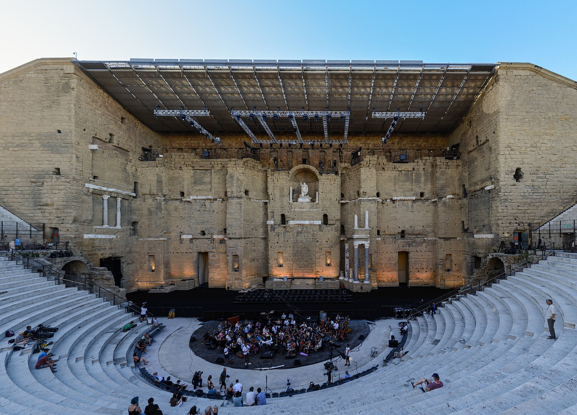 Le théâtre antique d’Orange vu des gradins : haut mur de scène de pierre percé de niches, statue impériale dans la niche centrale, gradins en hémicycle ; installations modernes de spectacle au-dessus de la scène