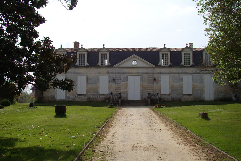 Allée de gravier conduisant à un logis noble modeste de plain-pied : longue façade claire à porte centrale sous fronton, lucarnes en toiture, volets fermés, pelouses et arbres