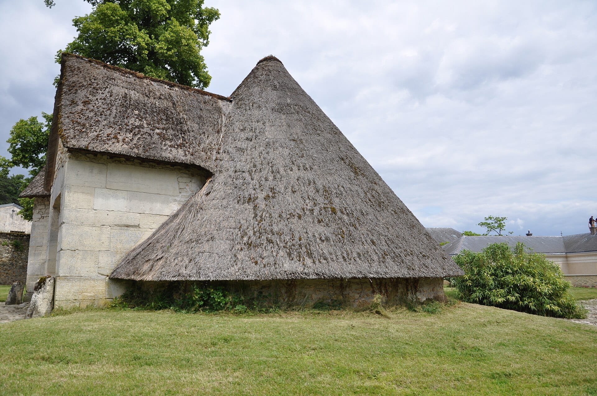 Une glacière du Petit Trianon vue de côté en été : vaste cône de chaume moussu descendant presque jusqu’au sol, accès de pierre de taille sur le flanc gauche, dans un cadre de gazon et d’arbres.