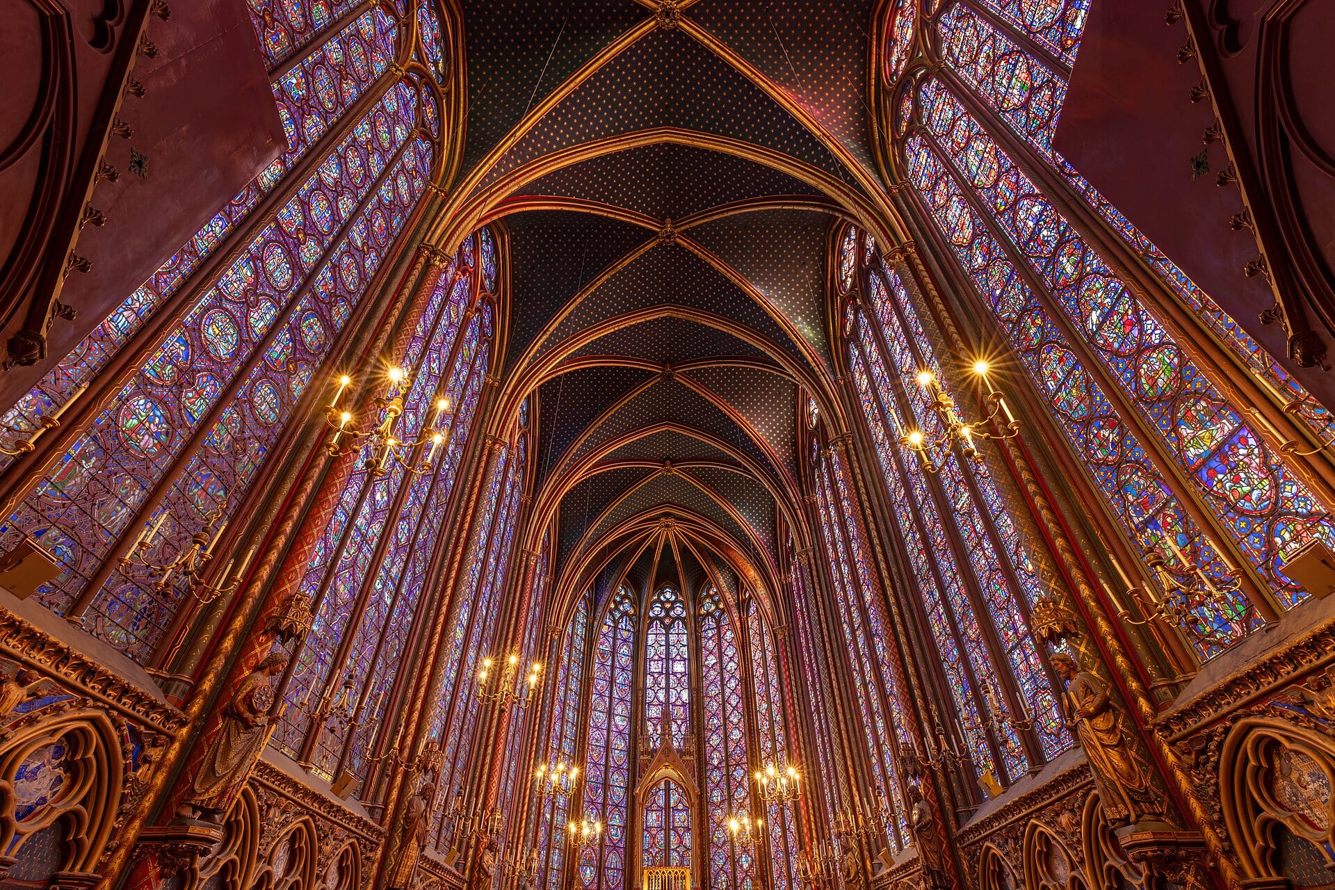 Intérieur de la haute chapelle de la Sainte-Chapelle de Paris : hautes verrières colorées séparées de fins meneaux, voûte sur croisée d’ogives étoilée bleu et or, lustres allumés