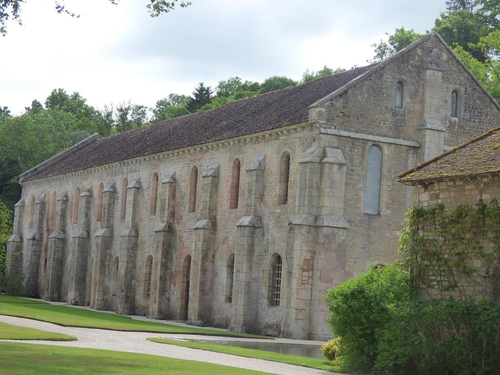 Longue halle de pierre à deux niveaux rythmée par une file de contreforts à ressauts, percée de fenêtres en plein cintre et en lancette, coiffée d’un toit de tuiles couvert de mousse et terminée par un mur-pignon.