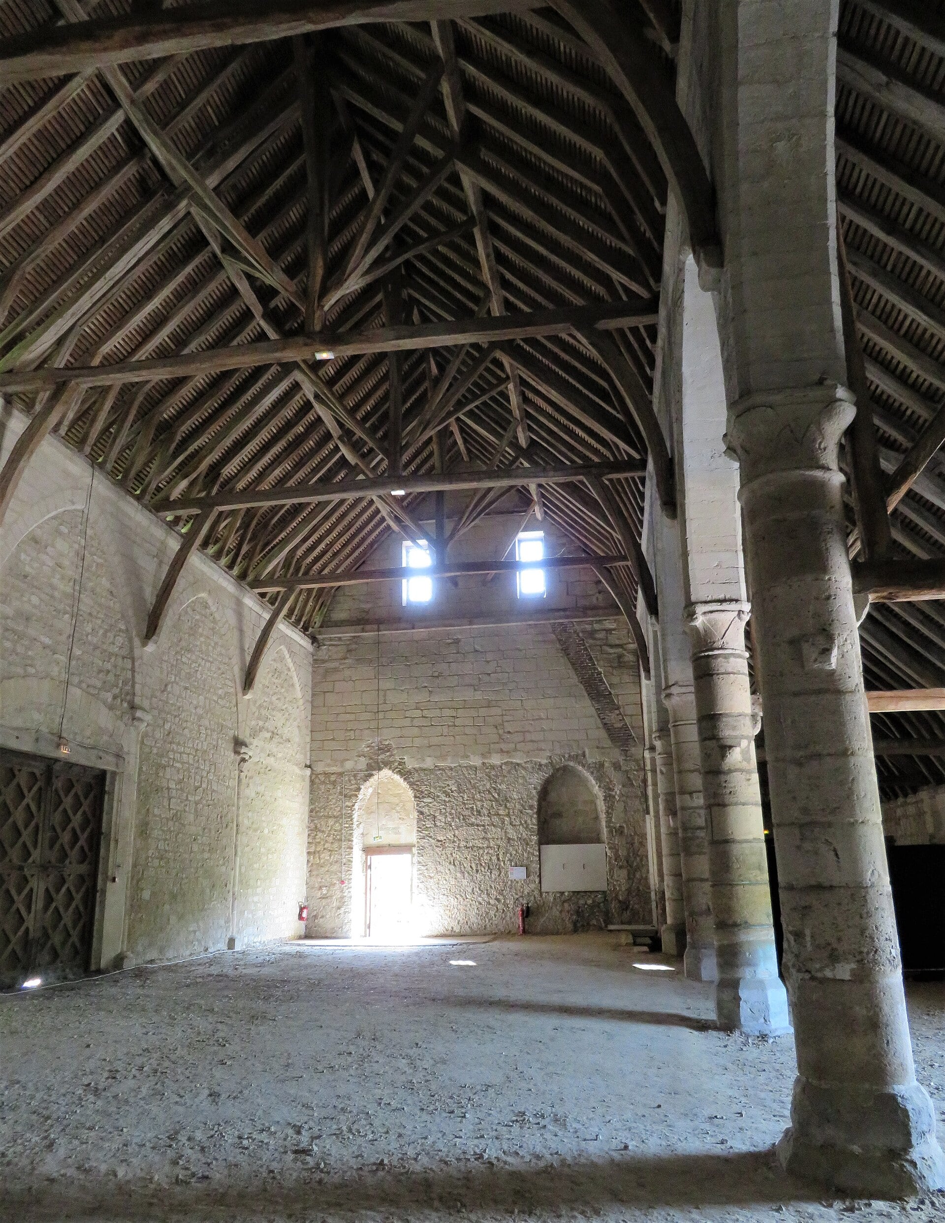 Intérieur d’une grange médiévale : haute charpente de bois à chevrons apparents, colonnes de pierre à chapiteaux sculptés soutenant la nef, sol de terre battue.