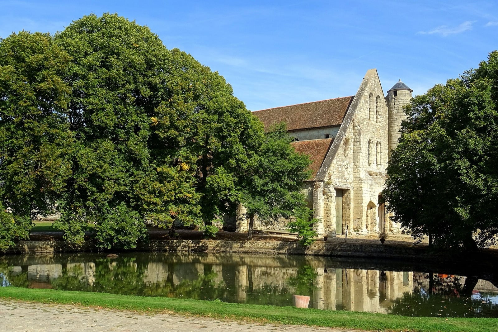 Grange de pierre claire vue depuis l’autre rive d’une mare : haut pignon à baies en lancette et tour ronde à droite, long toit de tuiles couvrant le vaisseau qui s’enfonce derrière un rideau d’arbres, le tout reflété dans l’eau.
