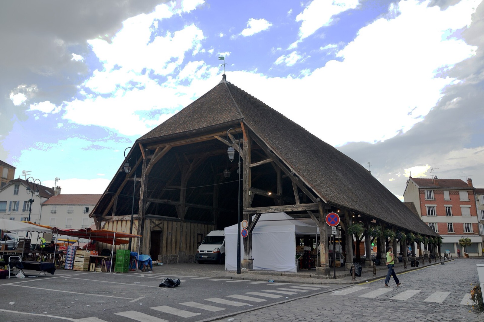 La halle d’Arpajon vue de trois quarts, poteaux de bois sous la longue toiture d’ardoise, étals de marché installés dessous