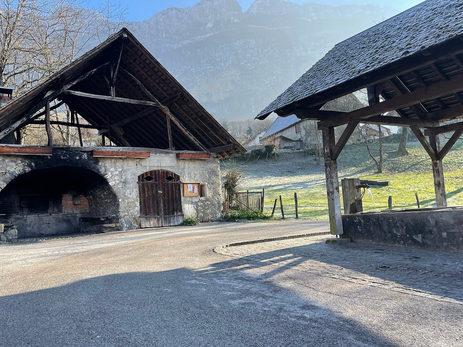Le four à pain communal d’un hameau savoyard, voûte de pierre et gueule noircie, face à une fontaine-lavoir sous son toit de bois