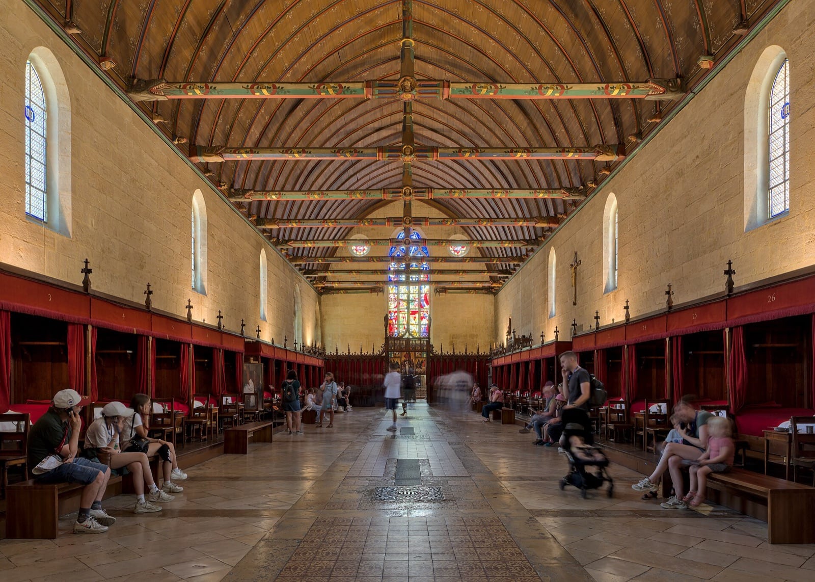 Grande salle des pauvres de l’hôtel-Dieu de Beaune : longue nef à voûte de bois lambrissée, deux rangées de lits à courtines rouges le long des murs, chapelle vitrée au fond ; des visiteurs sont assis et circulent.