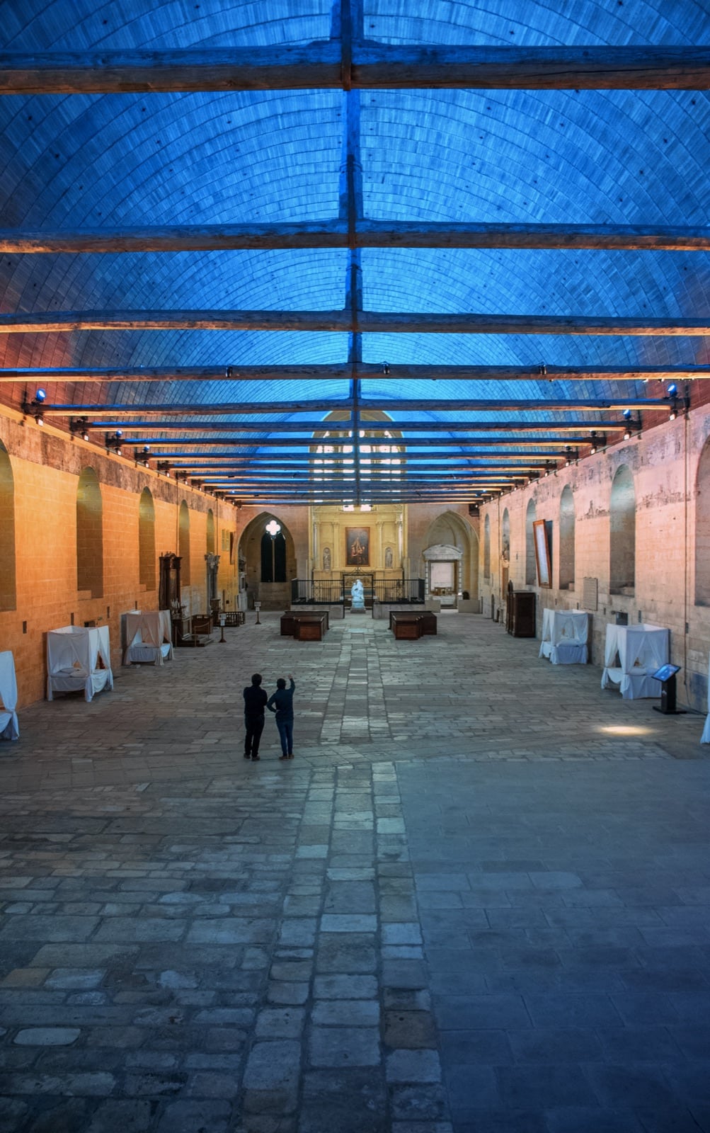 Grande salle des malades de l’hôtel-Dieu de Tonnerre : très longue nef unique couverte d’une voûte de bois en carène, murs de pierre à niches, sol pavé, chapelle au fond ; le lambris est baigné d’un éclairage bleu.