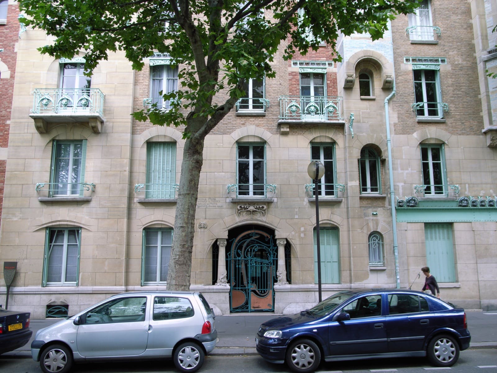 Façade du Castel Béranger à Paris : porte d’entrée fermée d’une grille en fer forgé aux courbes Art nouveau, murs mêlant pierre de taille et brique, balcons de fonte à motifs de masques