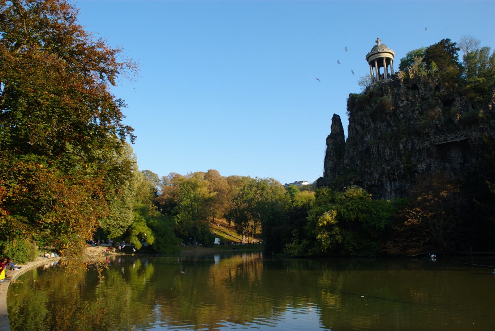 Le lac du parc des Buttes-Chaumont à Paris, dominé par l’île rocheuse escarpée que couronne le temple de la Sibylle, entre rochers artificiels et végétation d’automne.