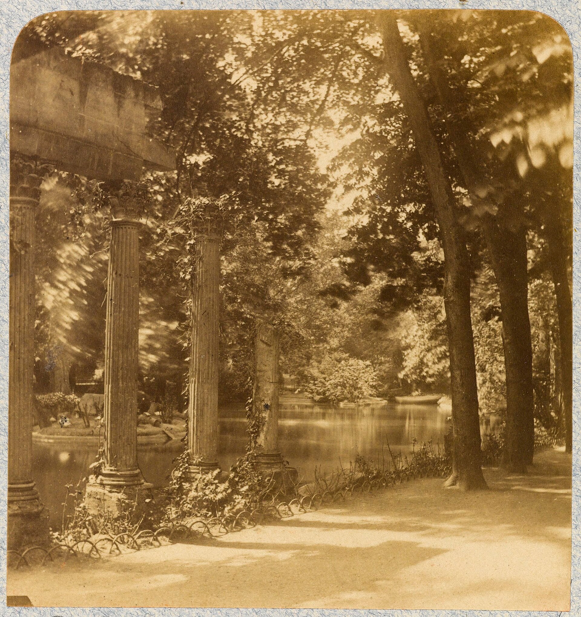 Photographie ancienne : la colonnade de la naumachie du parc Monceau à Paris, colonnes corinthiennes se reflétant dans le bassin, sous les arbres.