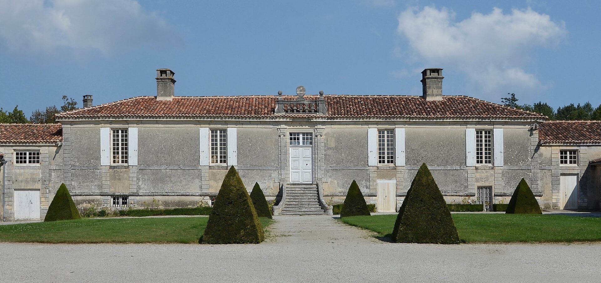 Longue façade classique d’un logis de plain-pied en calcaire clair : ordonnance symétrique, porte centrale à encadrement mouluré desservie par un perron, fenêtres à volets blancs, toiture de tuiles à faible pente, cheminées, ifs taillés en cône devant la maison