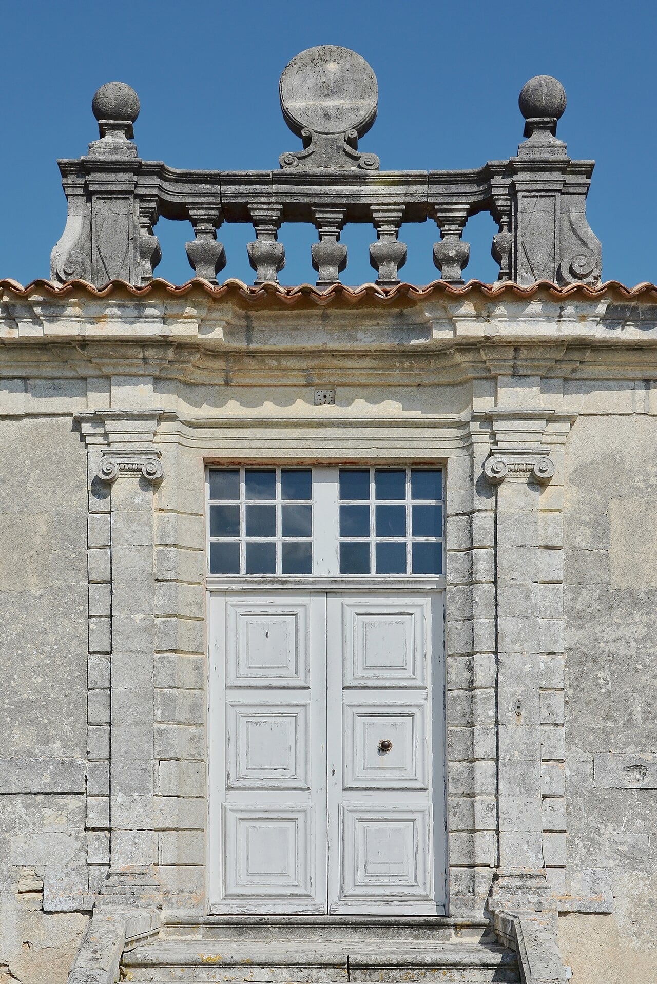 Gros plan de la travée centrale d’un logis classique : porte à deux vantaux blancs à panneaux surmontée d’une imposte vitrée, encadrement de pierre à pilastres ioniques et corniche moulurée, couronné d’une balustrade de pierre à balustres tournés portant un cadran solaire circulaire entre deux boules
