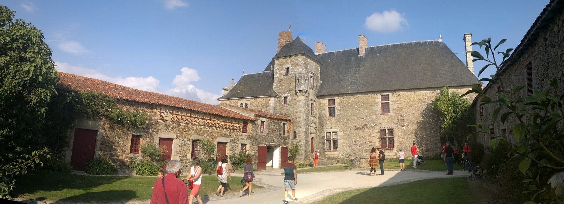 Cour fermée d’un ensemble seigneurial : à gauche de longs communs bas en moellon à portes rouges et toit de tuile ; au centre et à droite le logis à haute toiture d’ardoise, flanqué d’une tour d’escalier carrée, percé de fenêtres à croisée ; des visiteurs traversent la cour