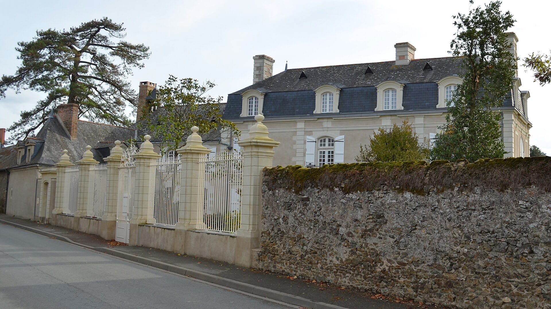 Vue depuis la rue d’un logis à façade claire et haute toiture d’ardoise à lucarnes cintrées et hautes cheminées de brique, précédé d’un haut mur de clôture en moellon et d’un portail monumental à piliers de pierre à boules et grille de fer blanche, communs bas sur la gauche