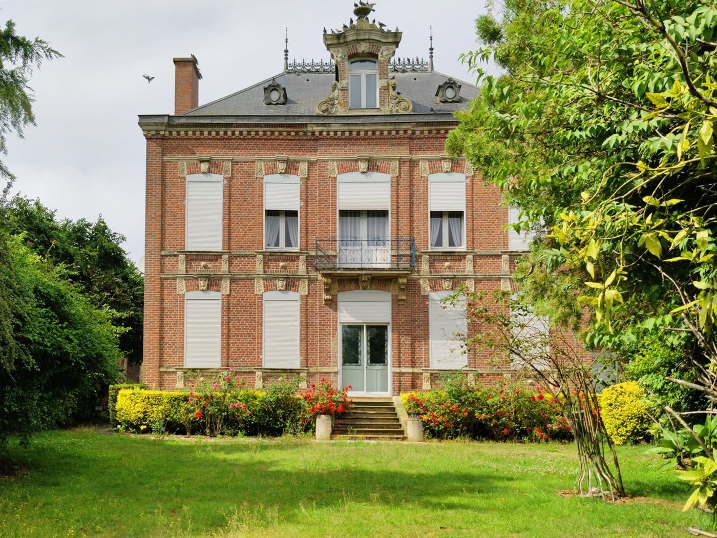 Maison de maître en brique rouge à décor de pierre claire, lucarne à volutes surmontée d’une urne, balcon en ferronnerie et jardin fleuri, à Contay.
