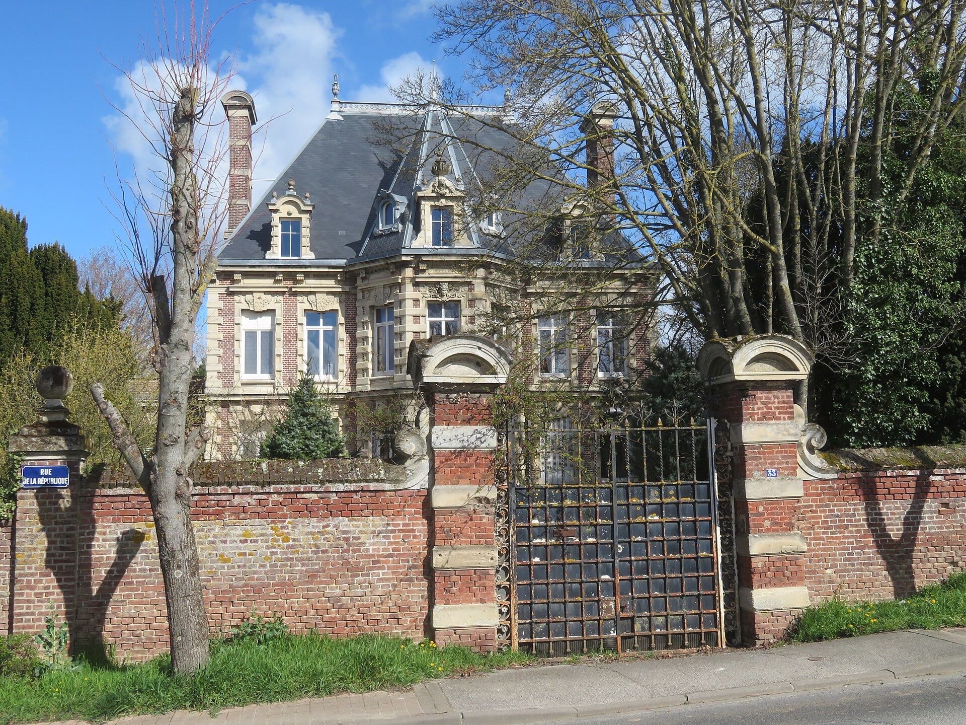 Maison de maître en brique et pierre à haut toit d’ardoise et lucarnes sculptées, vue depuis la rue par-dessus un mur de brique fermé d’une grille de fer entre piliers