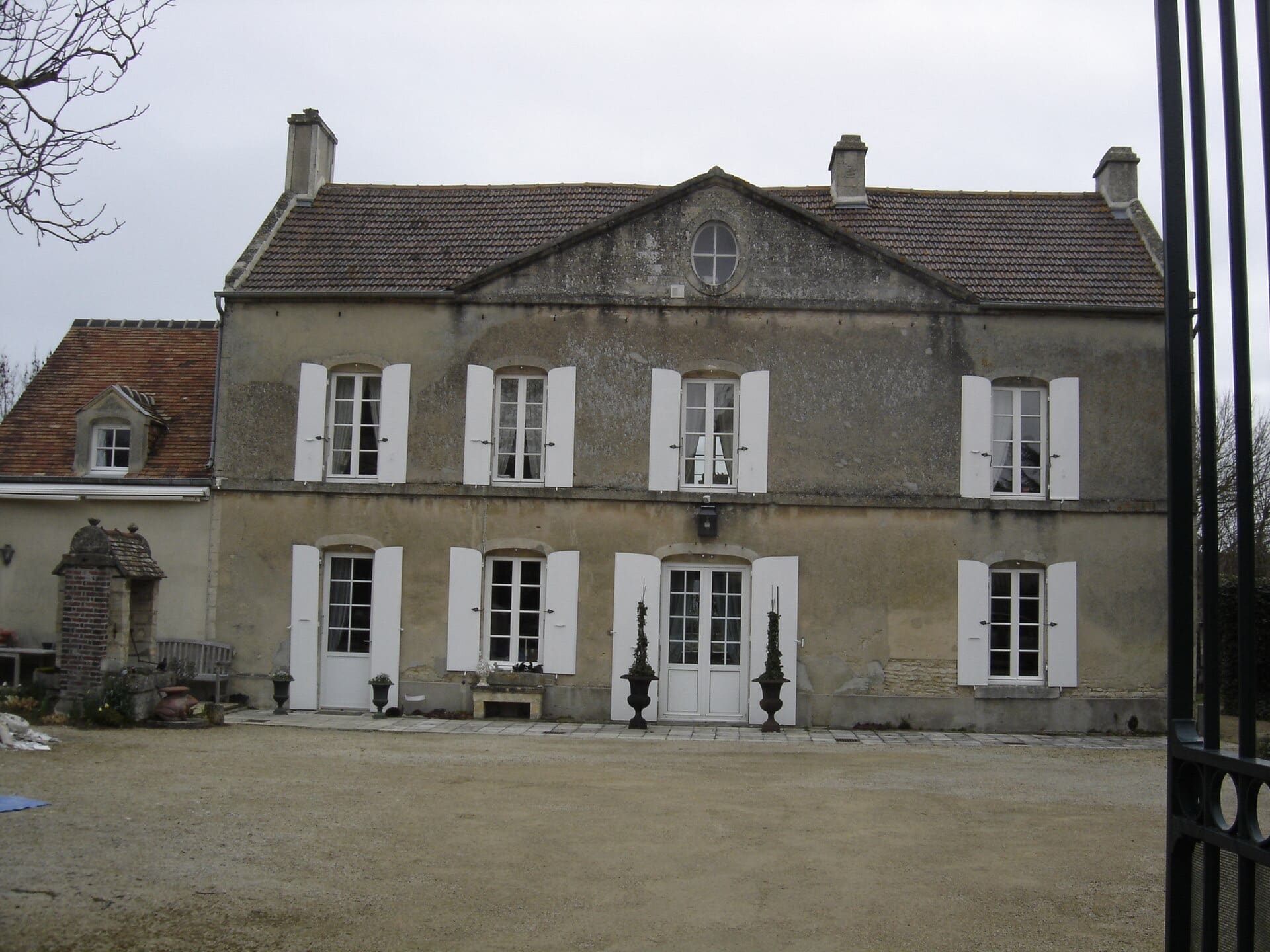 Façade enduite d’une maison de maître : quatre travées de fenêtres à volets blancs, deux portes-fenêtres centrales, fronton triangulaire percé d’un œil-de-bœuf au sommet du toit