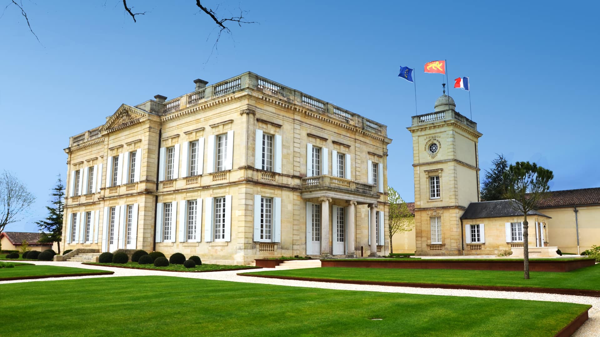 Château viticole en pierre blonde à avant-corps couronné d’un fronton et de pilastres colossaux, balustrade en couronnement, tour d’angle isolée portant des drapeaux, pelouse et allée
