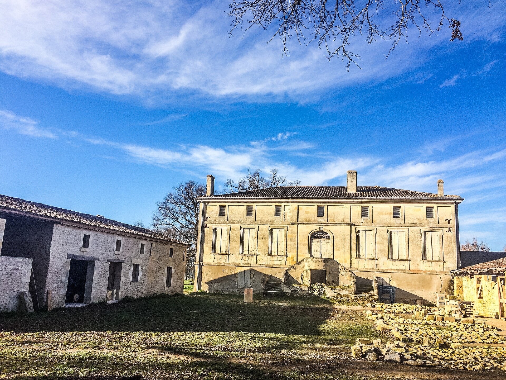 Maison de maître en pierre à rez-de-chaussée surélevé sur cellier, perron à double volée ruiné, façade symétrique à volets, flanquée à gauche d’un long commun bas en pierre