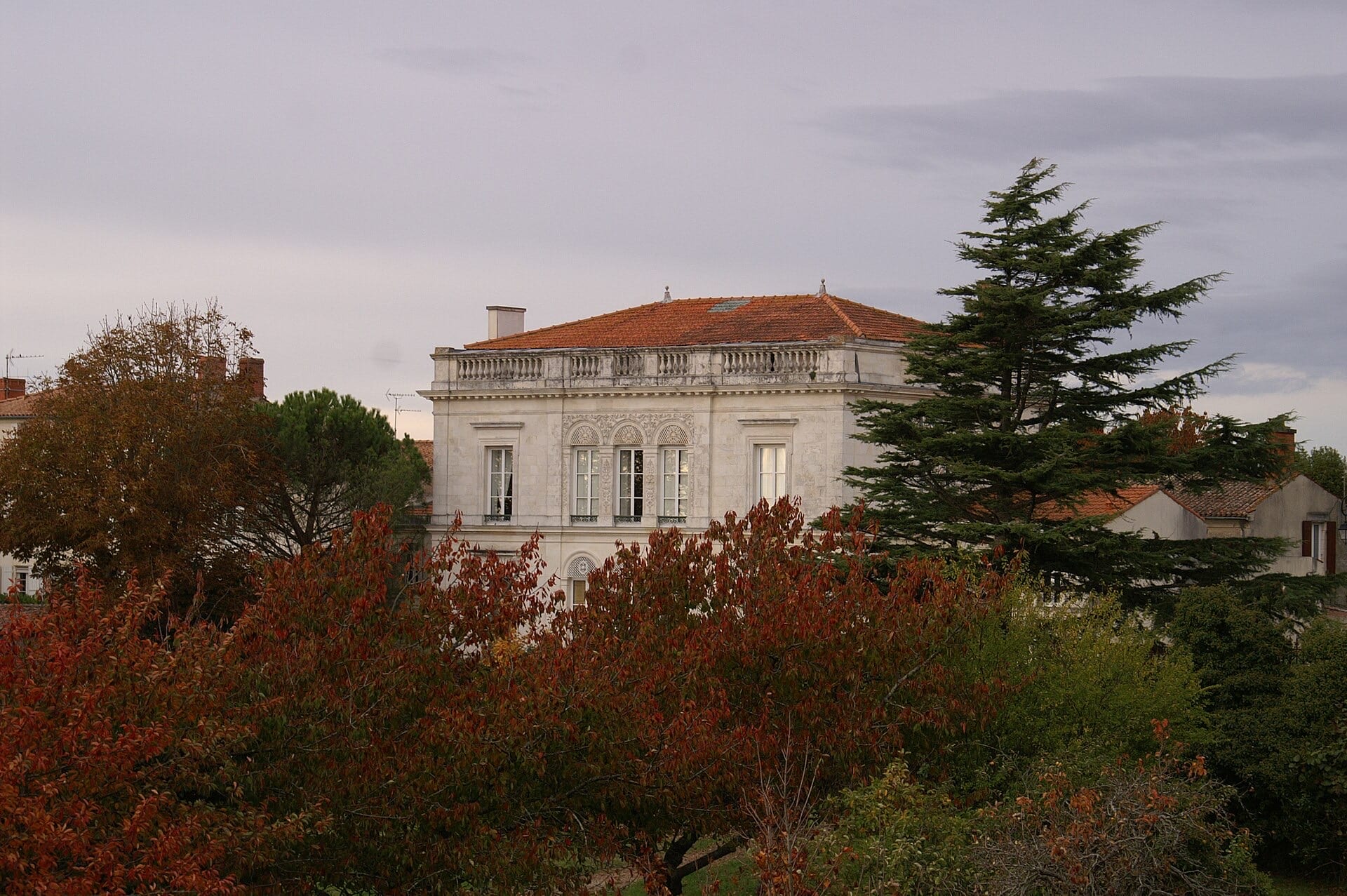 Maison de maître en pierre calcaire claire à toit couvert d’une balustrade, travée centrale richement sculptée d’arcatures, vue au-dessus d’arbres d’automne