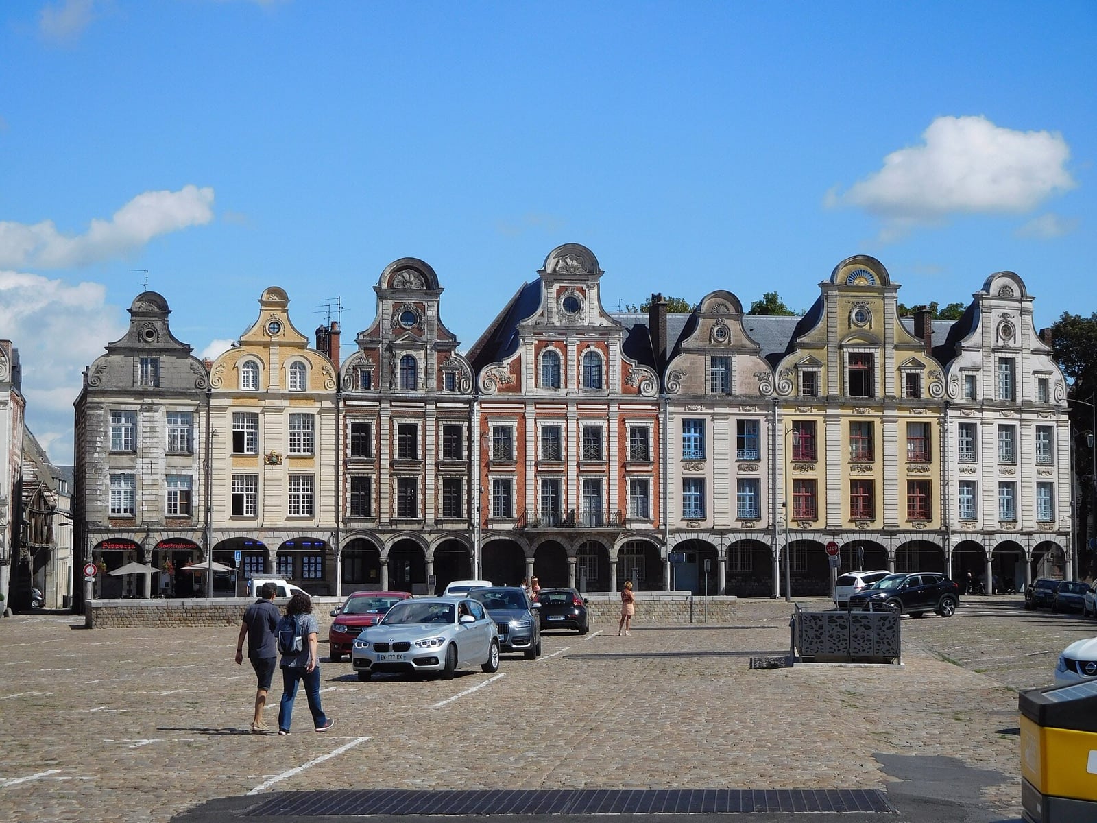 Rangée de maisons de la Grand-Place d’Arras à pignons courbes à volutes, façades de pierre et de brique, sur une galerie continue d’arcades au rez-de-chaussée.