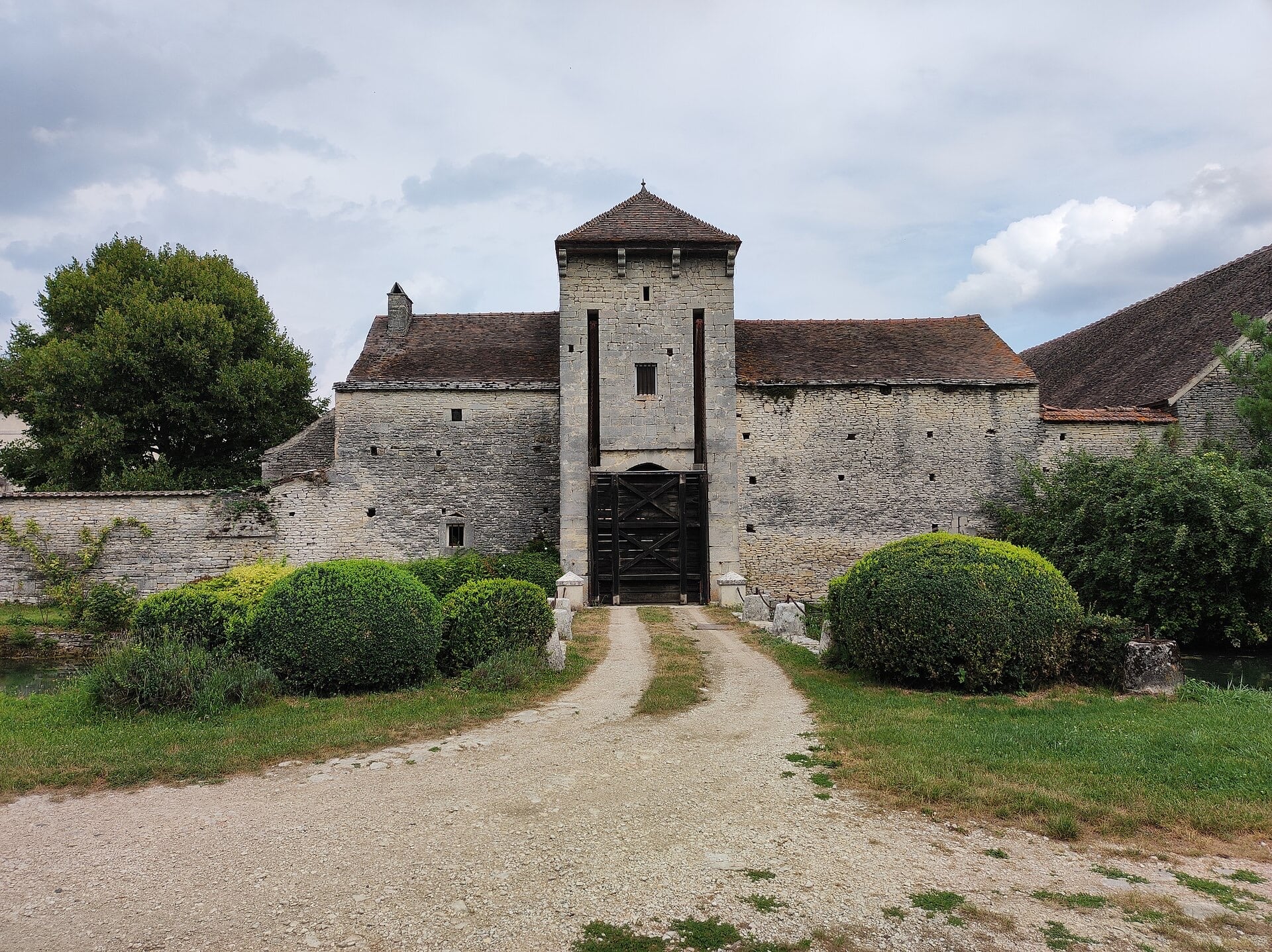 Vue frontale d’un châtelet d’entrée : haute tour-porte de pierre à toit de tuiles, porte charretière à vantaux de bois, allée gravillonnée franchissant le fossé