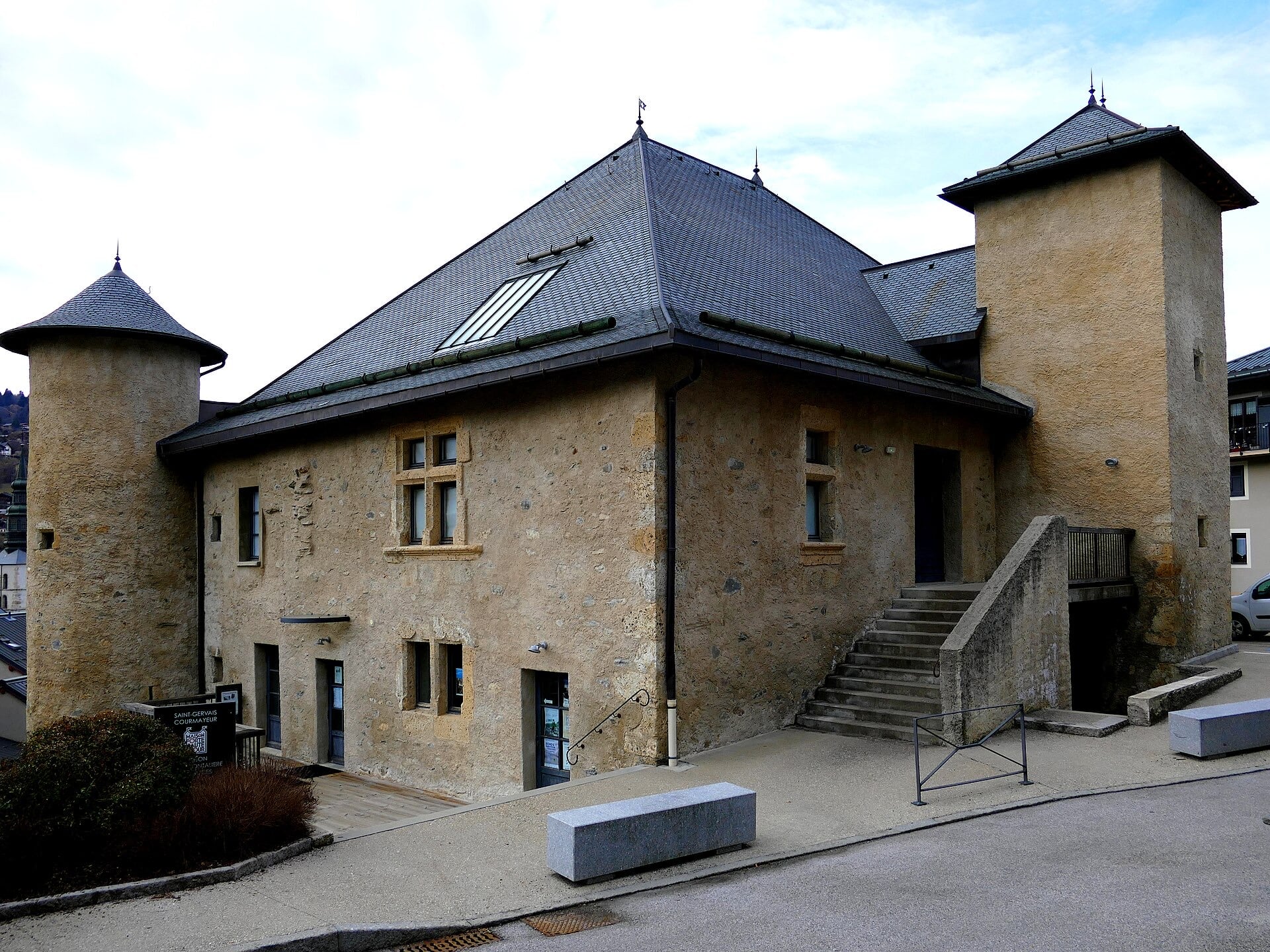 Logis de pierre de deux étages à haute toiture d’ardoise en croupe, flanqué d’une tour ronde coiffée d’un toit conique à gauche et d’une tour carrée à droite, percé de fenêtres à croisée de pierre, dans un environnement urbain