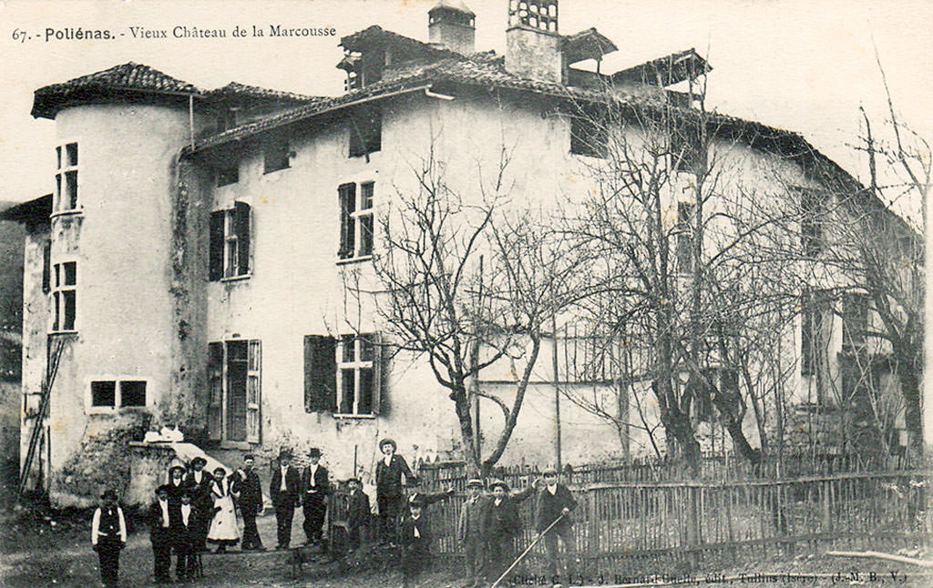 Carte postale ancienne en noir et blanc : logis rural blanchi à toiture irrégulière et hautes cheminées, flanqué d’une tour ronde arasée, aux fenêtres à volets, devant lequel pose un groupe de villageois en habits de 1900 derrière une clôture de bois