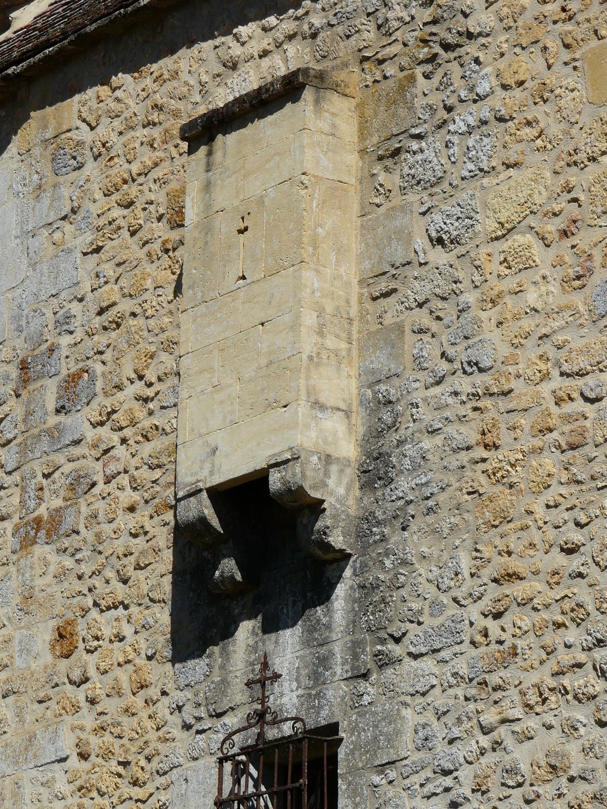 Détail d’un mur de pierre : bretèche de pierre en saillie sur corbeaux, percée d’une fente de tir verticale et ouverte par en dessous d’un mâchicoulis, dominant une baie fermée d’une grille de fer forgé