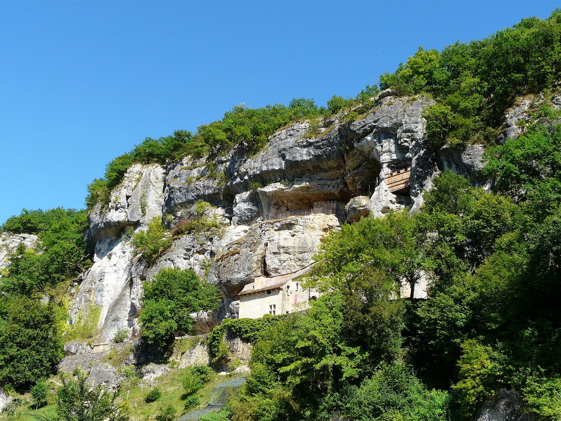 Contre une haute falaise calcaire dominant une vallée boisée, un logis de pierre à toit de tuiles blotti sous le surplomb rocheux, surmonté de galeries de bois logées dans des abris-sous-roche