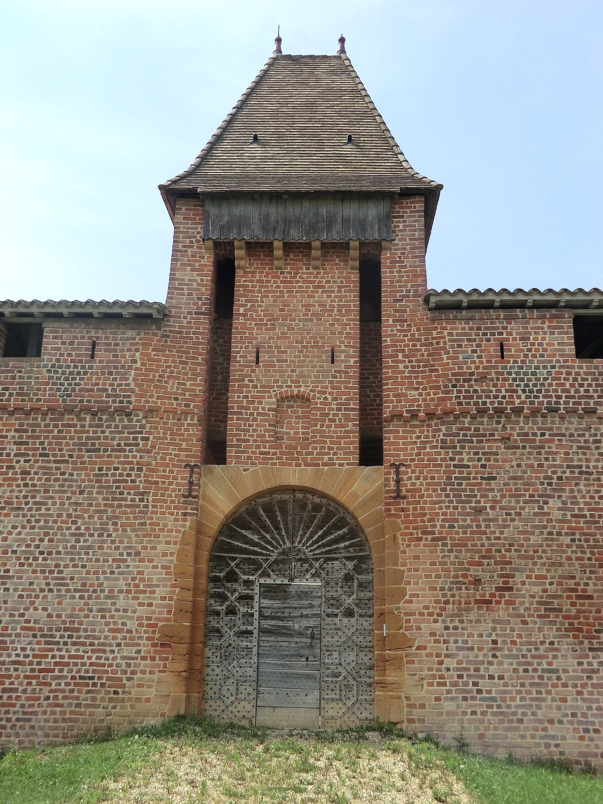 Tour-porte de brique rouge crénelée coiffée d’un toit de tuiles en pavillon, surmontant un hourd de bois en encorbellement ; en dessous, une grande porte charretière à vantaux de bois cloutés sous un arc de pierre dorée