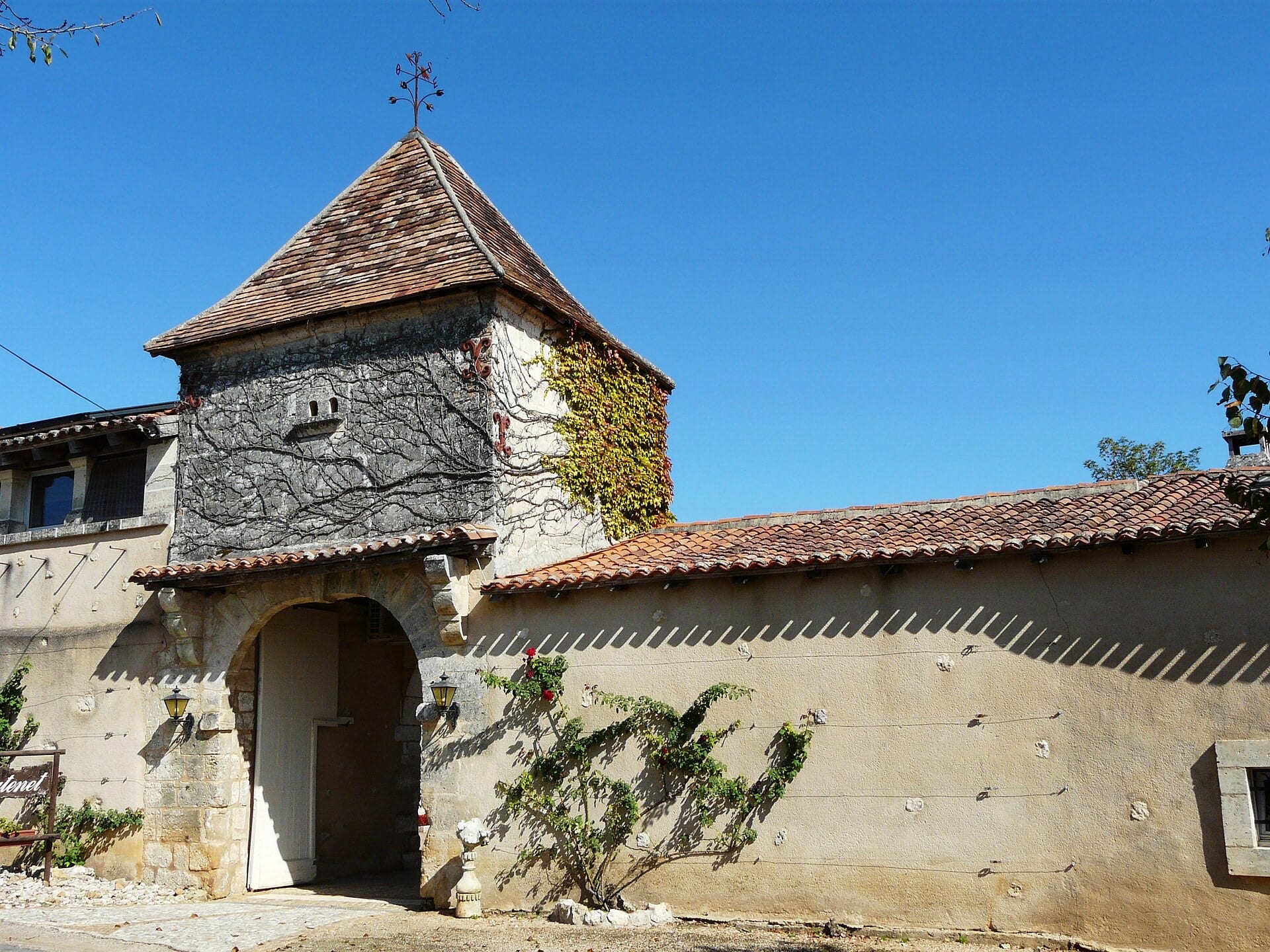 Porche d’entrée en arc de pierre de taille percé dans le mur de clôture, surmonté d’un pigeonnier carré à toit pyramidal de tuiles et girouette