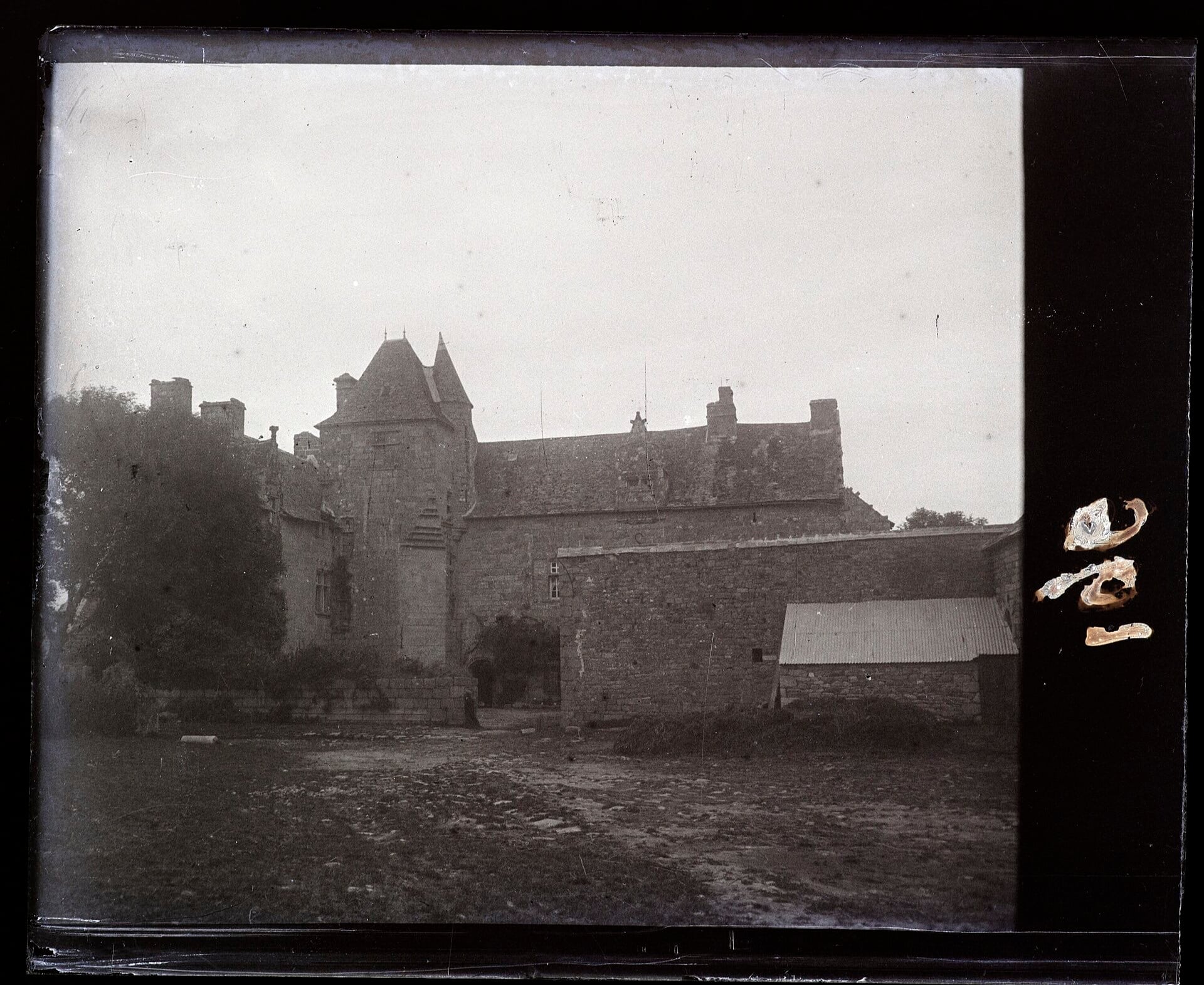 Photographie ancienne en noir et blanc d’un manoir de pierre à tour d’escalier polygonale et hautes cheminées, cour close et communs, émulsion abîmée sur un bord
