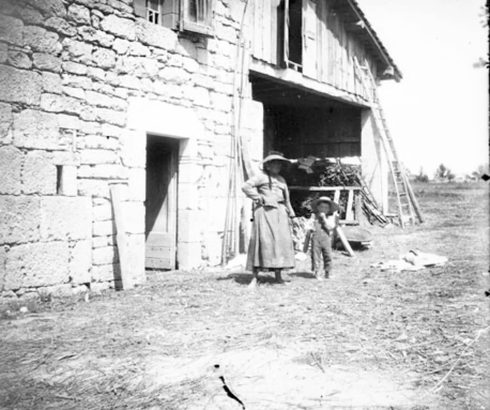 Photographie ancienne : une femme en long tablier et large chapeau de paille, une main sur la hanche, se tient près d’un enfant coiffé d’un chapeau devant une bâtisse rustique ; mur de pierre à encadrements de pierre de taille autour d’une porte basse, niveau supérieur bardé de planches avec baie ouverte de grange et échelle appuyée.