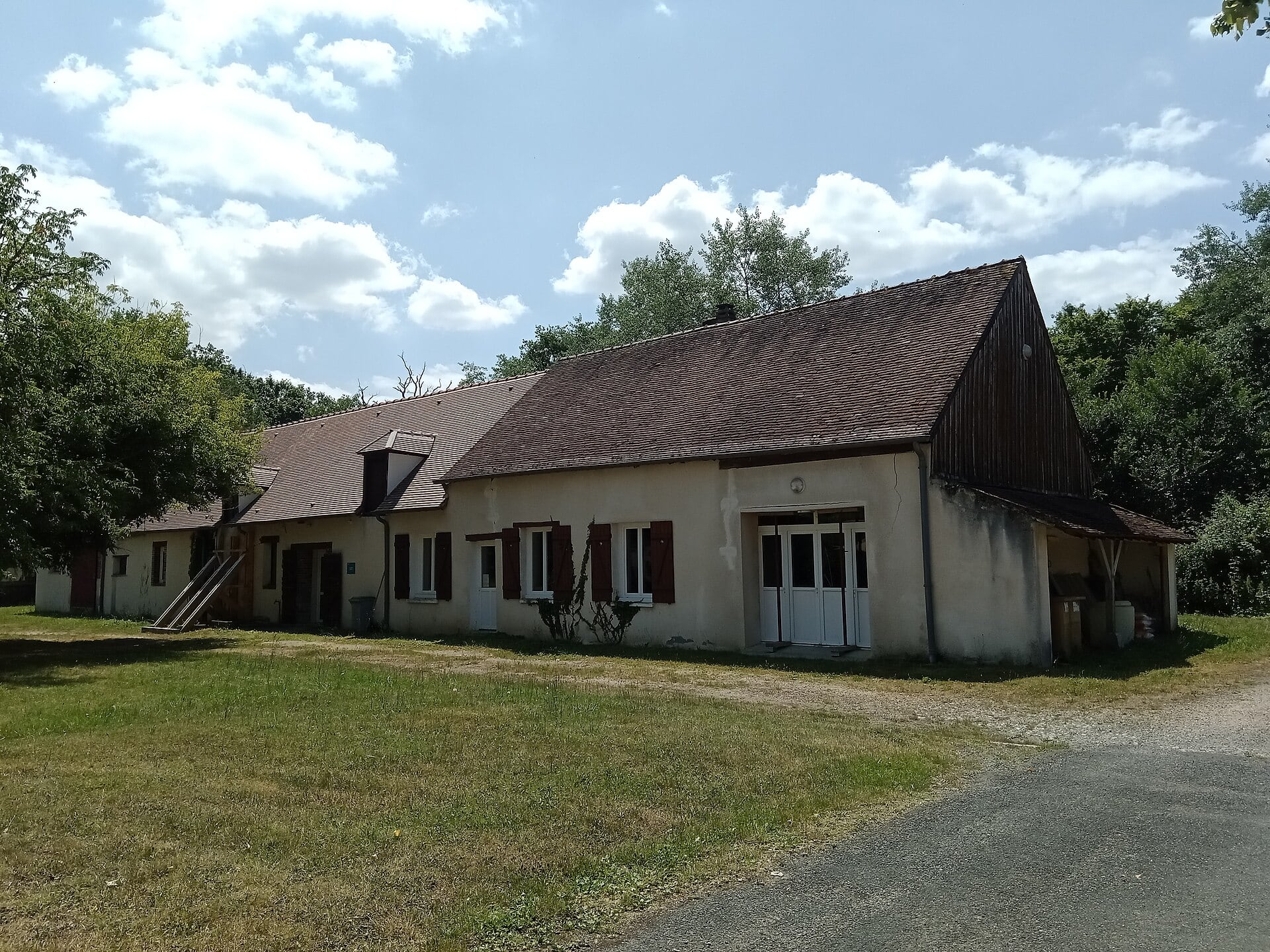 Long corps de logis bas en pierre enduite, sous toit de tuiles à forte pente, avec plusieurs travées de fenêtres à volets rouges et, à droite, une dépendance ouverte servant de grange ou d’étable.