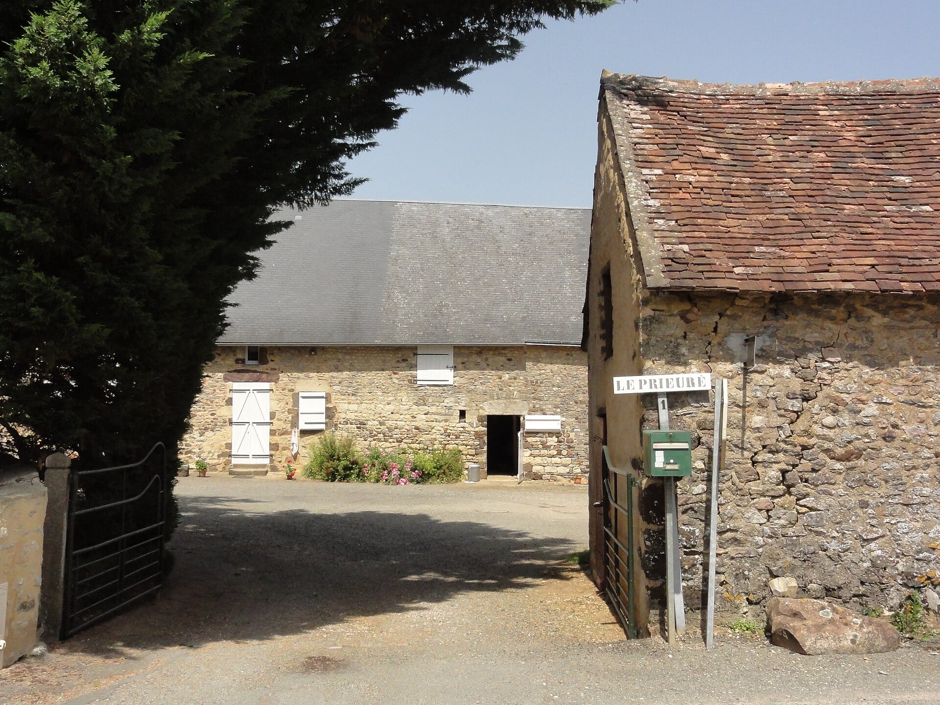 Corps de ferme en pierre calcaire à deux toitures, l’une en ardoise et l’autre en tuile, avec portail d’entrée et plaque murale portant l’inscription « Le Prieuré ».