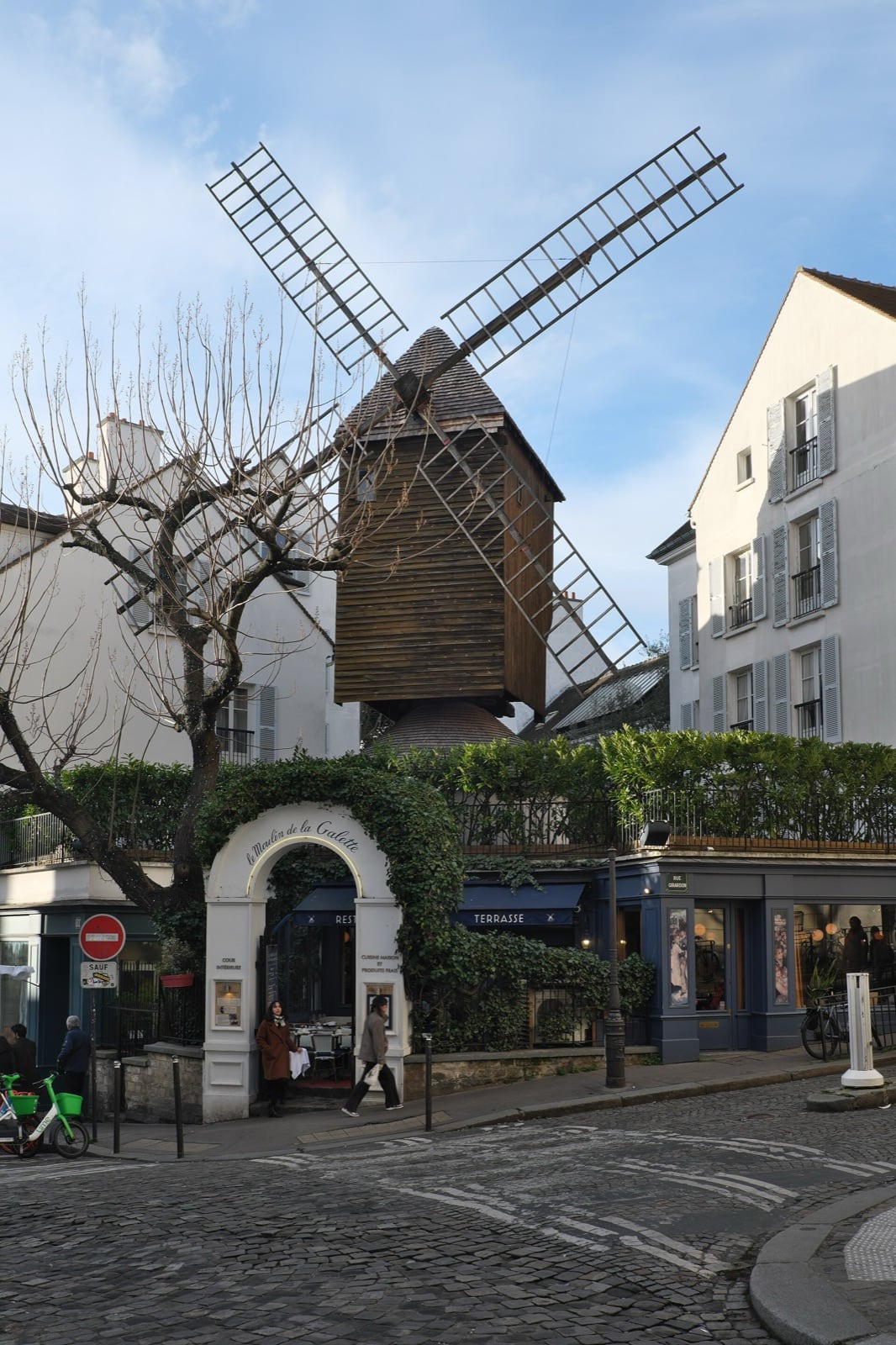 Moulin à vent de bois monté sur un socle maçonné, portant quatre ailes en châssis, dominant l’arche d’entrée d’un restaurant à l’angle d’une rue pavée.