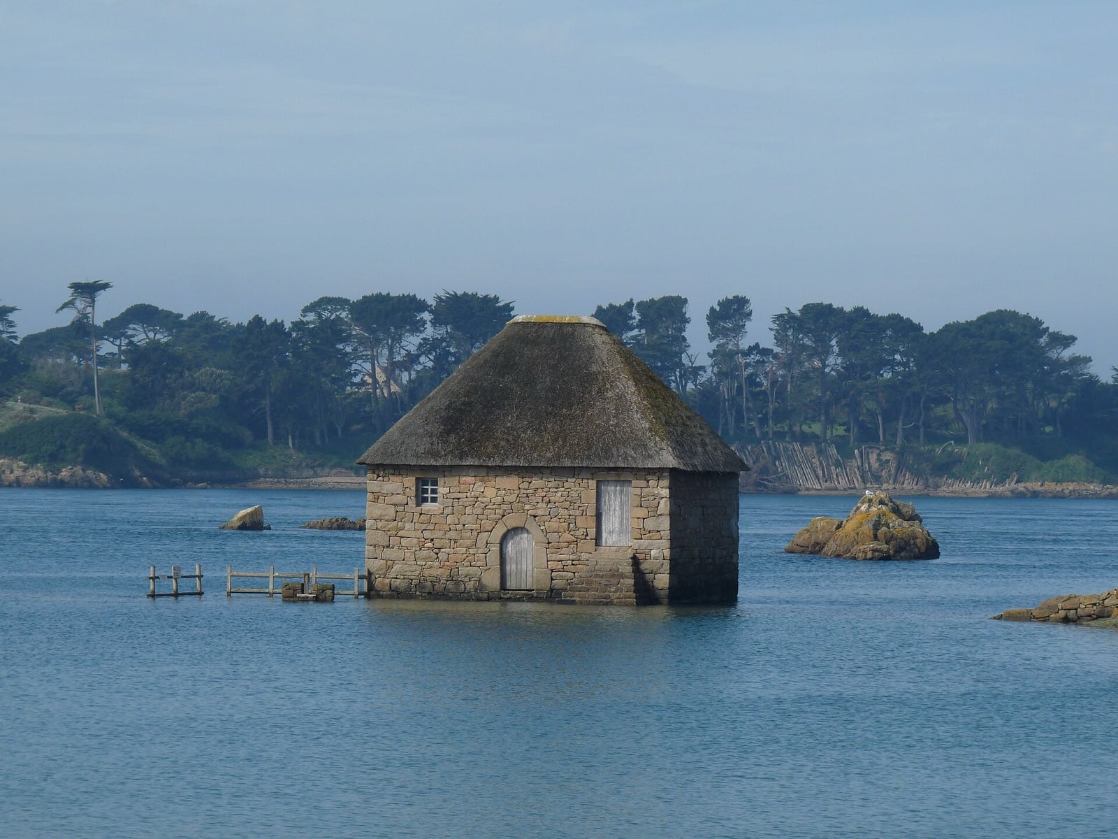Petit moulin de granit à toit conique de chaume, isolé au milieu de l’eau à marée haute, devant une rive boisée et des rochers.