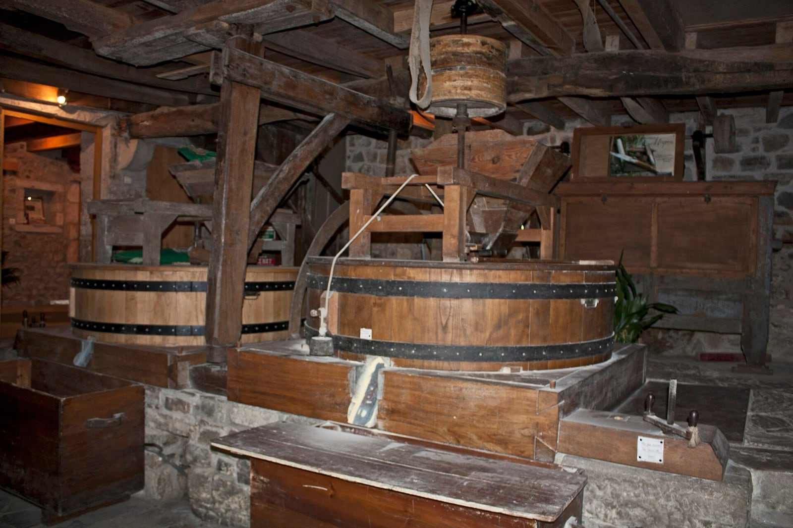 Intérieur de moulin : deux couples de meules enfermés chacun dans une archure de bois cerclée de fer, surmontés d’une trémie en bois, sous une charpente à poutres.