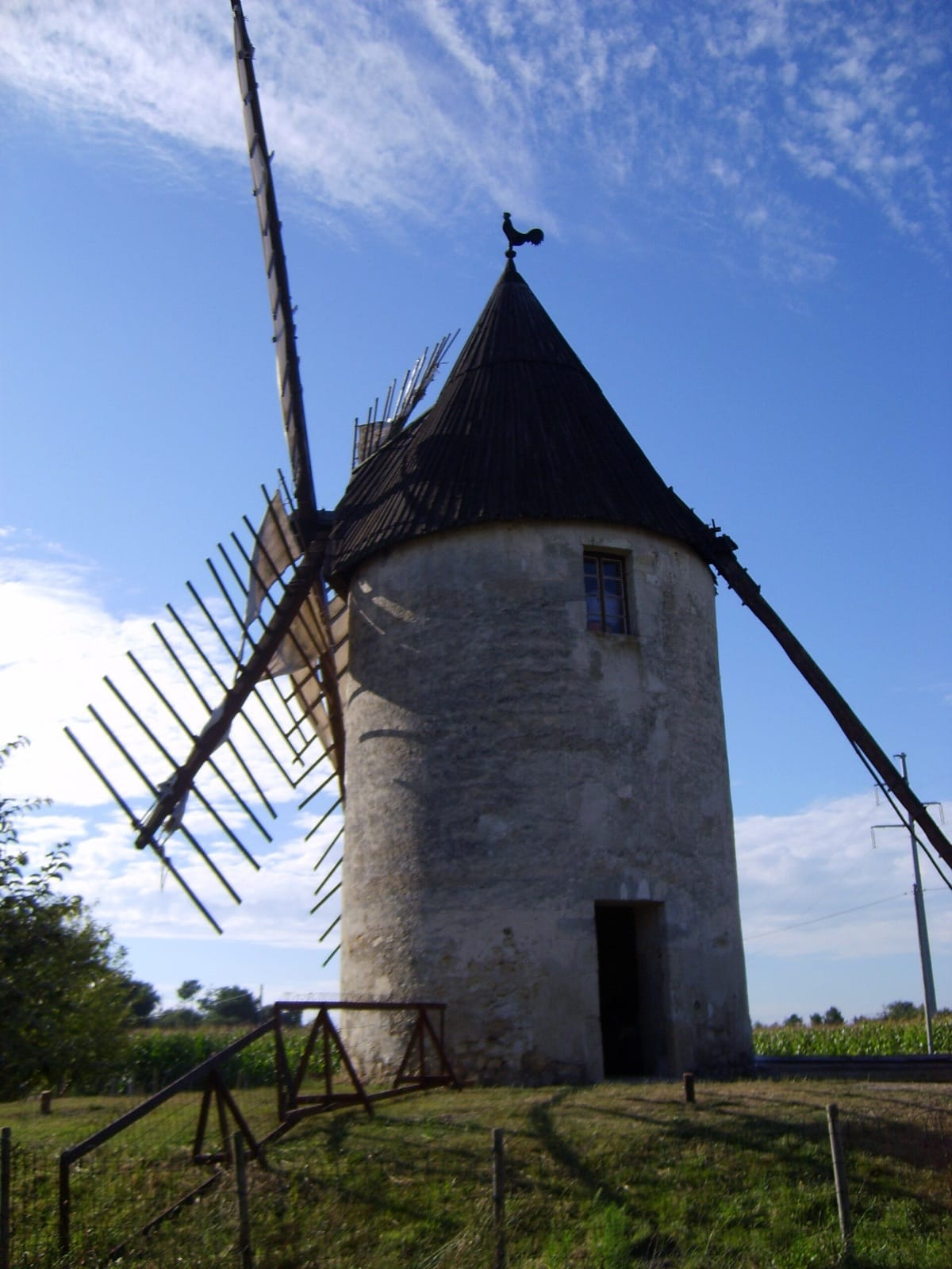 Moulin à vent en pierre : tour cylindrique blanchie coiffée d’une calotte conique en bardeaux surmontée d’un coq, portant les châssis d’ailes en bois nu.