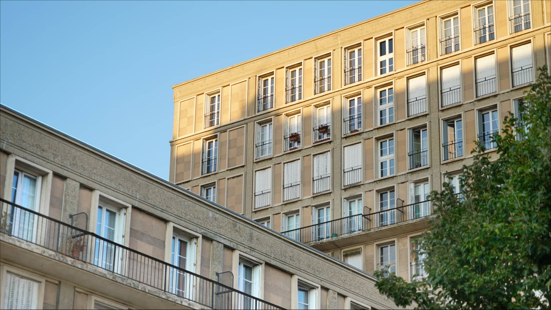 Façades d’immeubles de la reconstruction du Havre par Auguste Perret : trame régulière de béton, fenêtres alignées et balcons, en contre-plongée dans une lumière dorée