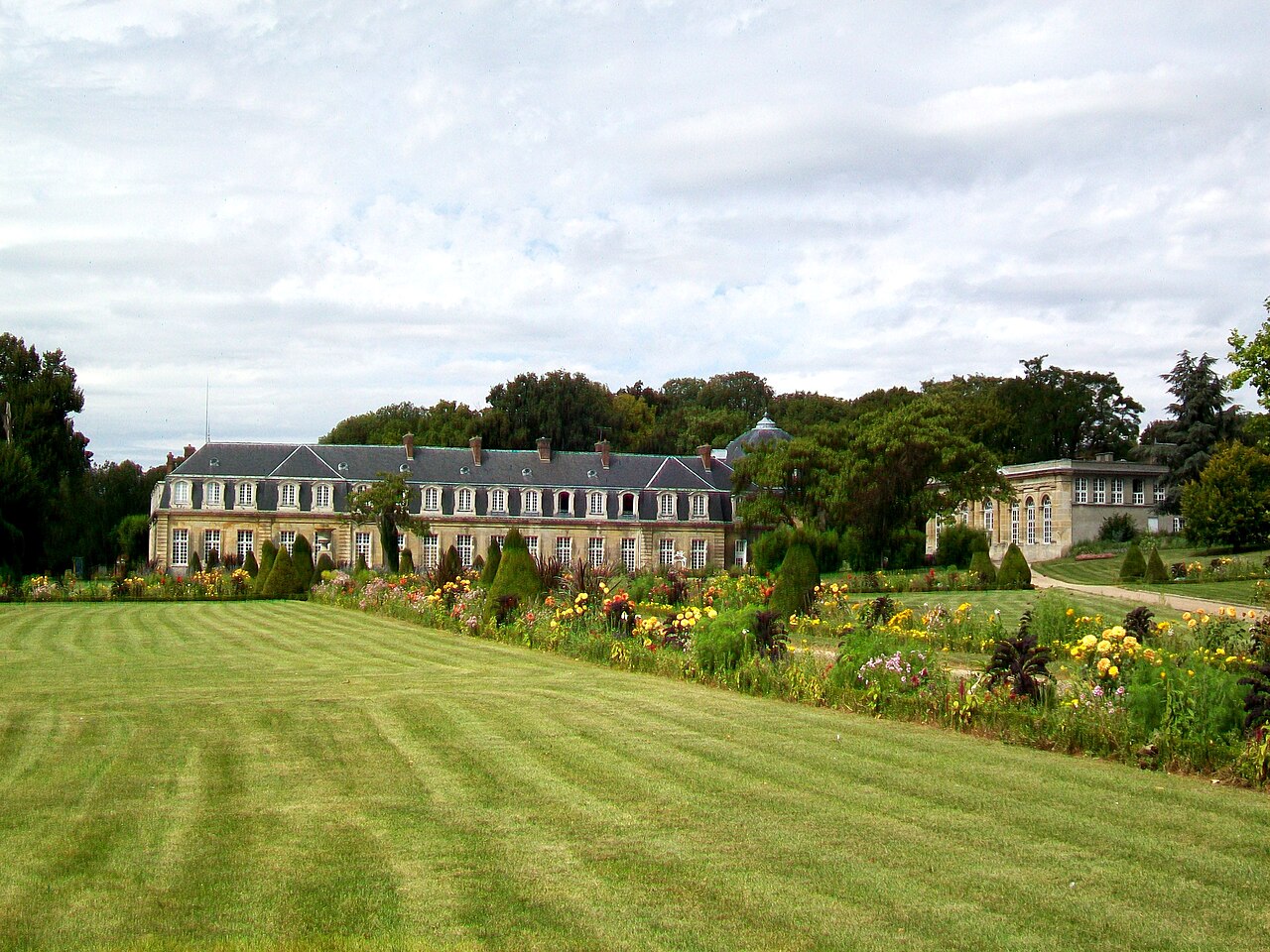 Le château d’Arnouville-lès-Gonesse (Val-d’Oise) et, à droite dans le parc, son orangerie basse à arcades vitrées, devant de grands massifs fleuris.