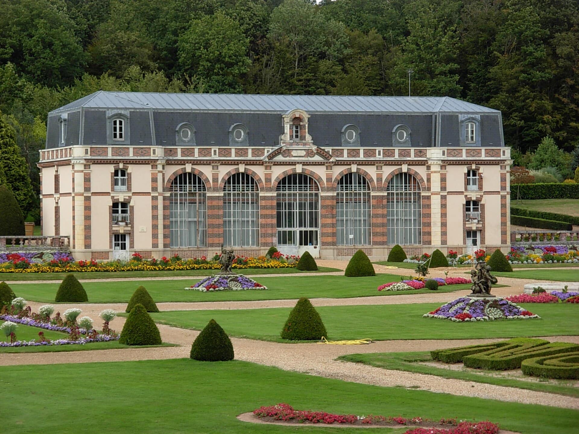 L’orangerie du château des Vaux (Eure-et-Loir), longue galerie à hautes baies cintrées vitrées en pierre et brique, ouverte au midi sur un jardin à parterres fleuris et ifs taillés.