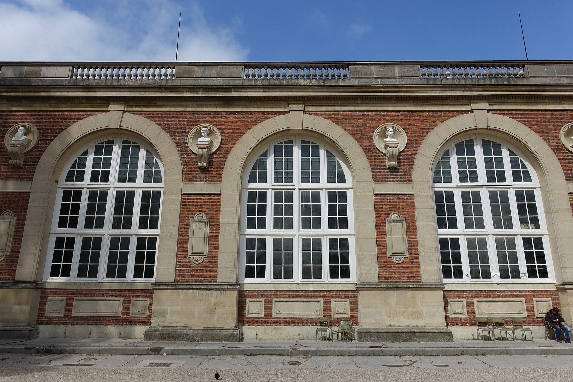 La façade de l’orangerie du jardin du Luxembourg : hautes baies cintrées, parement de brique et pierre, bustes en médaillon.