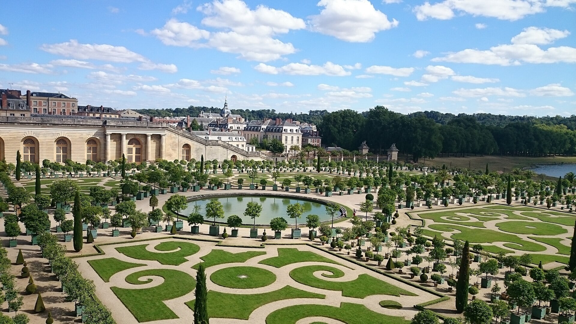 Le parterre bas de l’Orangerie de Versailles vu de la terrasse : orangers en caisses et broderies de gazon autour d’une pièce d’eau circulaire, la galerie à gauche.